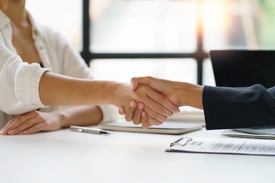 Deux professionnels en tenue business assis à une table de réunion dans un bureau moderne, examinant ensemble des documents contractuels et des dossiers d'appels d'offres. L'un pointe des clauses techniques sur un document, l'autre prend des notes. Environnement lumineux avec baies vitrées donnant vue sur un port ou contexte urbain. Atmosphère de concentration et d'expertise, documents et ordinateur portable visibles. Photographie professionnelle en lumière naturelle, ambiance sérieuse et méthodique.